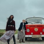 Two women in hippie outfits beside a vintage red van decorated with peace symbols