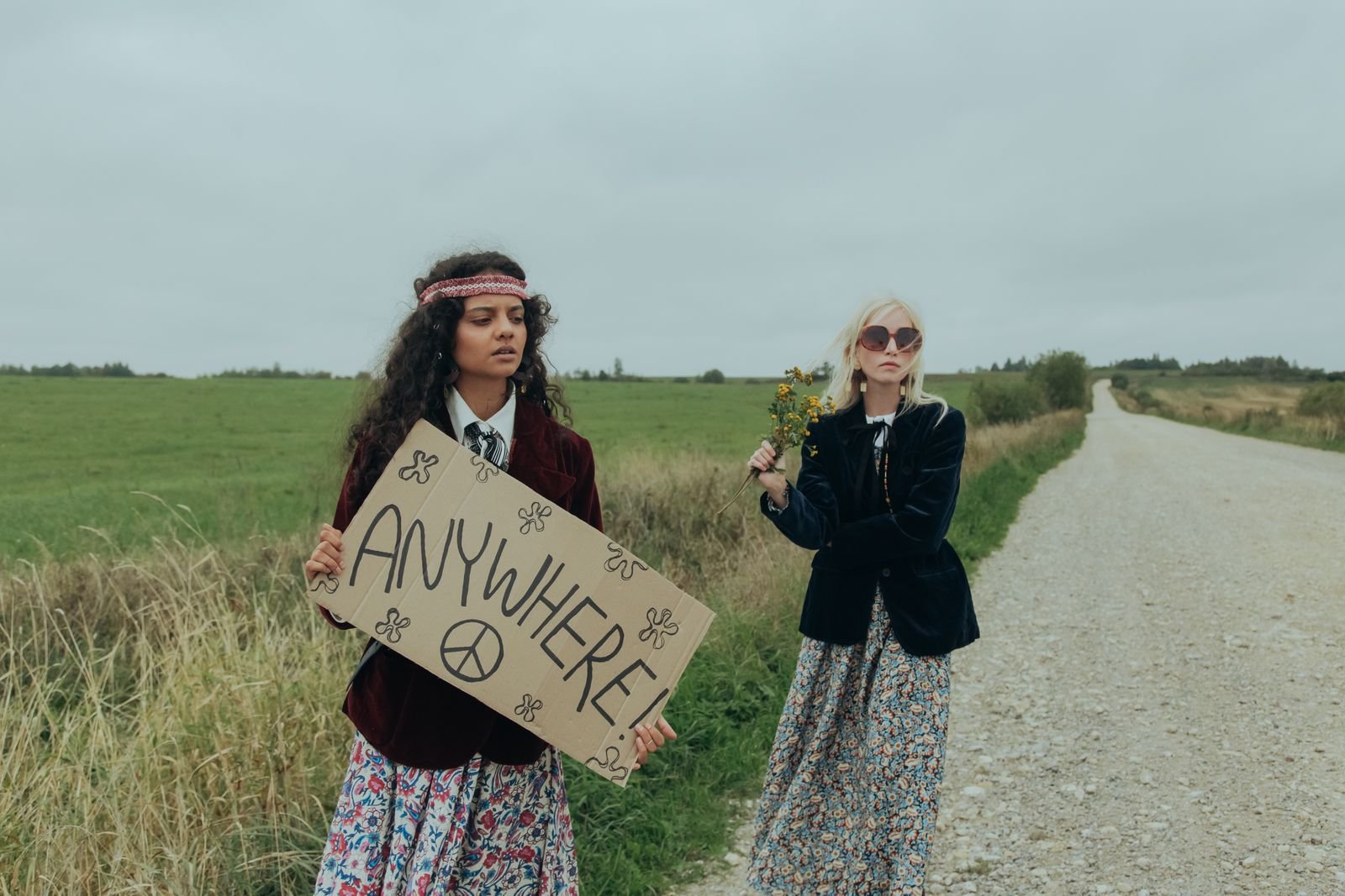 Two women in hippie-style clothes holding a hitchhiking sign on a rural road